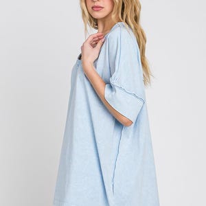May include: Light blue t-shirt with a relaxed fit and short sleeves. The shirt has a V-neck and visible stitching details. The model is wearing white shorts. Casual wear.