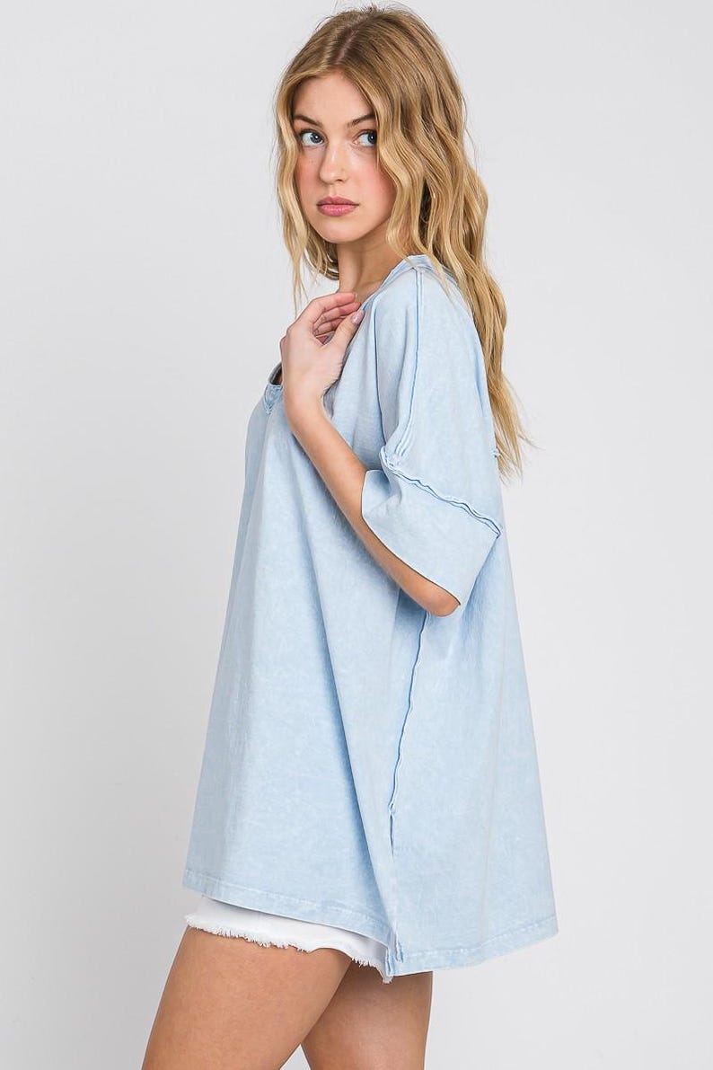 May include: Light blue t-shirt with a relaxed fit and short sleeves. The shirt has a V-neck and visible stitching details. The model is wearing white shorts. Casual wear.