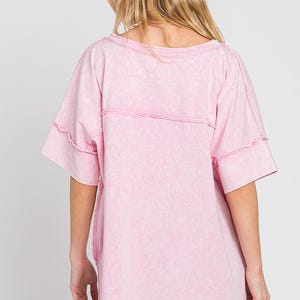 May include: A light pink, short-sleeved t-shirt with a washed effect. The shirt has a wide, round neckline and visible stitching details across the chest and sleeves. The garment is made of a soft, comfortable material.