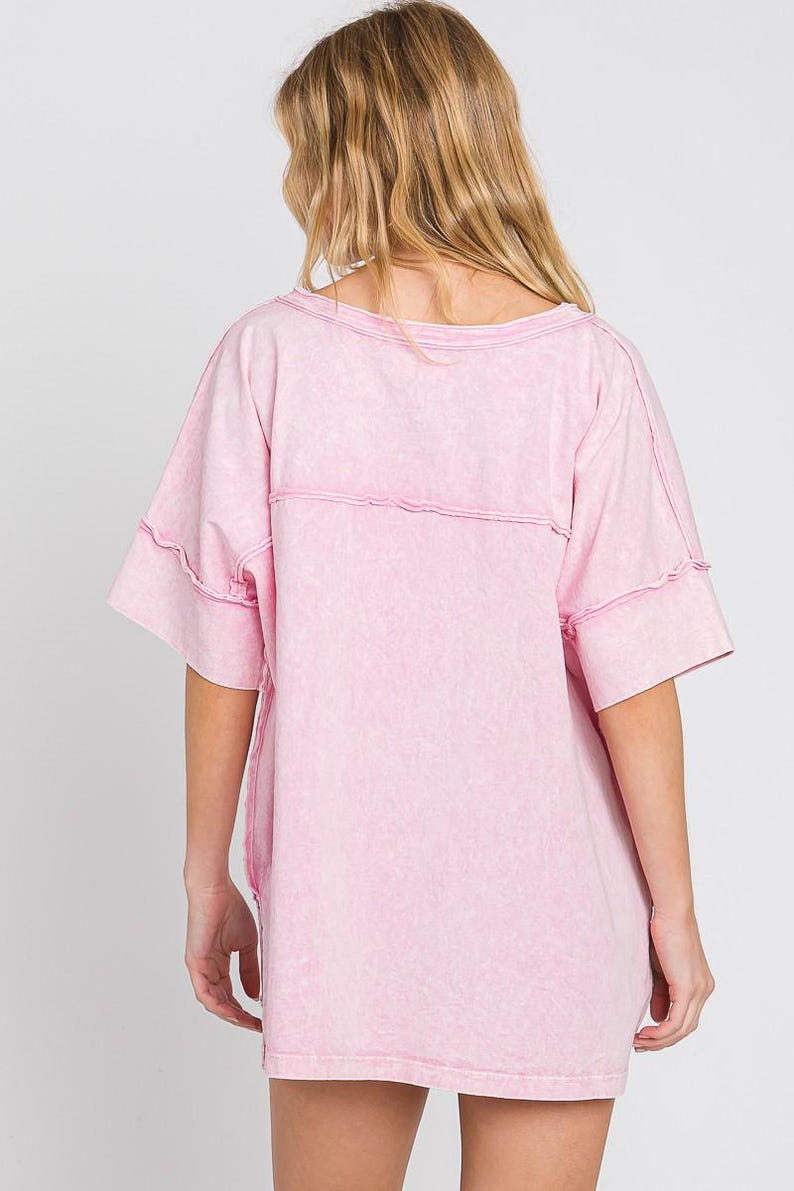 May include: A light pink, short-sleeved t-shirt with a washed effect. The shirt has a wide, round neckline and visible stitching details across the chest and sleeves. The garment is made of a soft, comfortable material.
