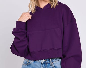 Puede incluir: Una sudadera corta de color morado intenso con capucha, mangas largas con puños acanalados, dobladillo acanalado a juego y un bolsillo canguro frontal grande. La prenda se combina con pantalones cortos de mezclilla azul claro con detalles desgastados y un botón plateado.