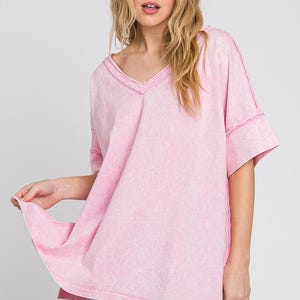 May include: A light pink, oversized V-neck t-shirt with short sleeves. The shirt has a washed, textured appearance and visible stitching along the seams. The model is wearing the shirt.