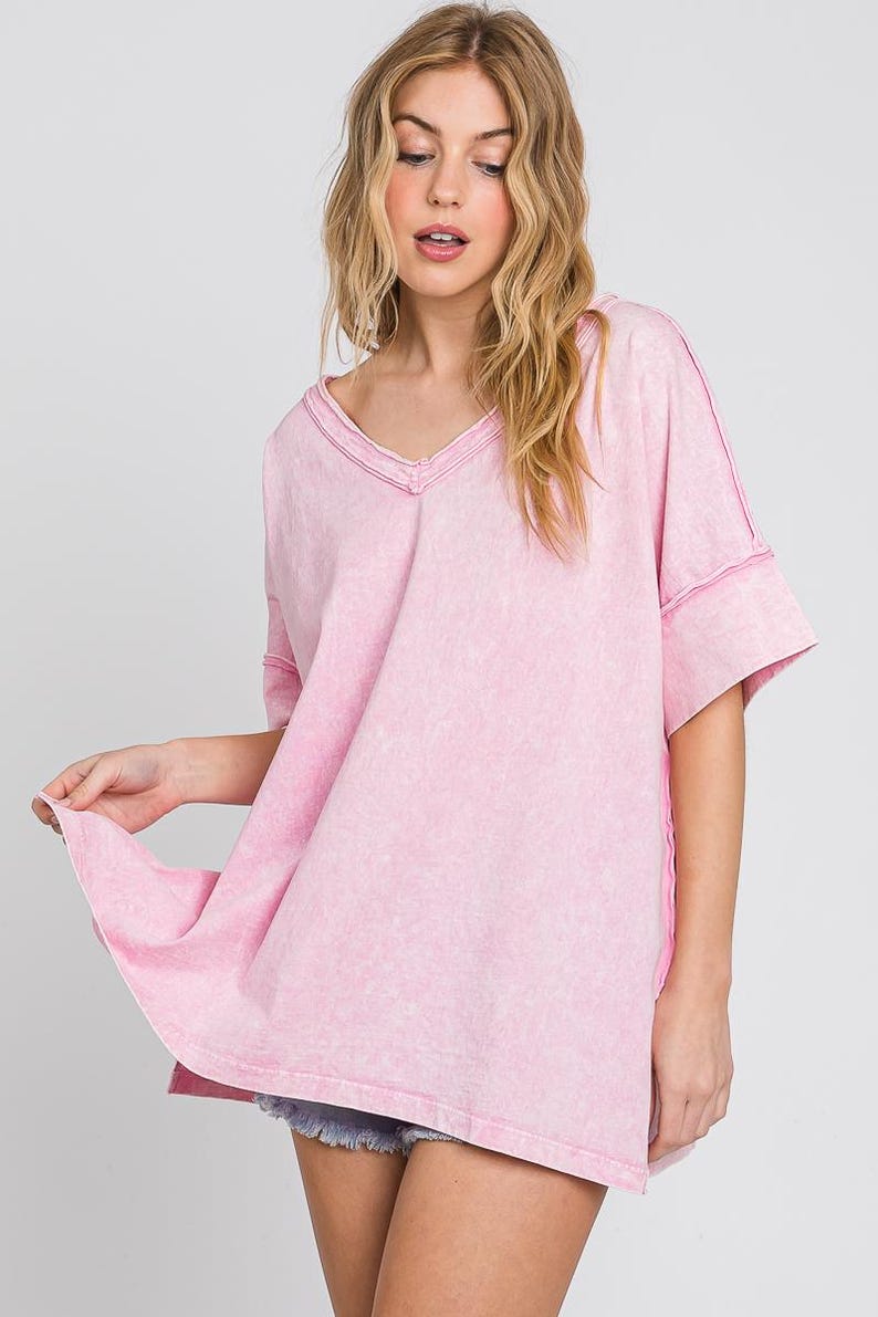 May include: A light pink, oversized V-neck t-shirt with short sleeves. The shirt has a washed, textured appearance and visible stitching along the seams. The model is wearing the shirt.