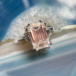 May include: A silver ring with a large, clear emerald-cut diamond. The diamond is set in a prong setting and has a slight pink hue. The ring is on a blue and white agate background.