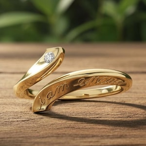 May include: A gold bypass ring featuring a single round clear gemstone set at one end and the cursive engraved text "i am enough" along the polished band. The ring has an open design with tapered ends and a smooth, reflective metallic finish.