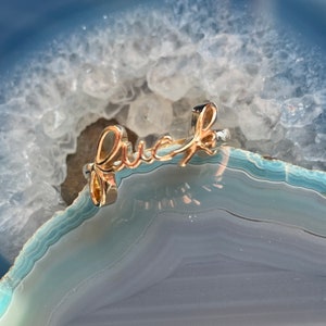 Puede incluir: Un anillo dorado con la palabra "Luck" en cursiva. El anillo se encuentra sobre un fondo azul y blanco, creando un contraste entre el metal y la piedra.