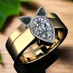 Può includere: Un anello di fidanzamento con un diamante a forma di pera e tre diamanti neri più piccoli incastonati in una fascia d'oro. L'anello è una doppia fascia con una fascia più larga nella parte inferiore.