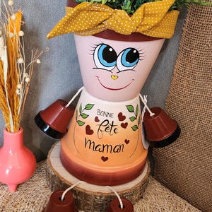 Peut inclure: Pot de fleur en terre cuite fantaisiste en forme de personne, avec des traits de visage peints, un bandeau jaune à pois et de faux bras et jambes. Le pot est rempli de verdure et les mots "Bonne Fête Maman" sont écrits sur le devant.