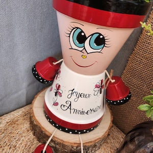 Peut inclure: Pot de fleur en terre cuite peint à la main en forme de poupée. Le pot présente un visage souriant avec de grands yeux bleus, un bord rouge et noir, et le texte français "Joyeux Anniversaire". Des cloches rouges servent de bras et de jambes.