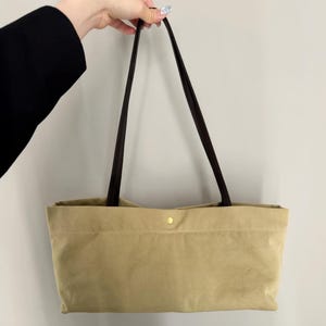 Può includere: Una borsa tote rettangolare di colore beige chiaro con una consistenza liscia e opaca. La borsa presenta due lunghi e sottili manici a spalla marrone scuro e un singolo piccolo bottone a pressione in metallo dorato al centro del bordo.