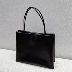 Op de afbeelding: Een zwarte draagtas met twee dunne, gebogen handvatten. De tas heeft een rechthoekige vorm met een platte bodem en een gladde, glanzende afwerking. De handvatten zijn ook zwart en lijken van hetzelfde materiaal te zijn als de tas.
