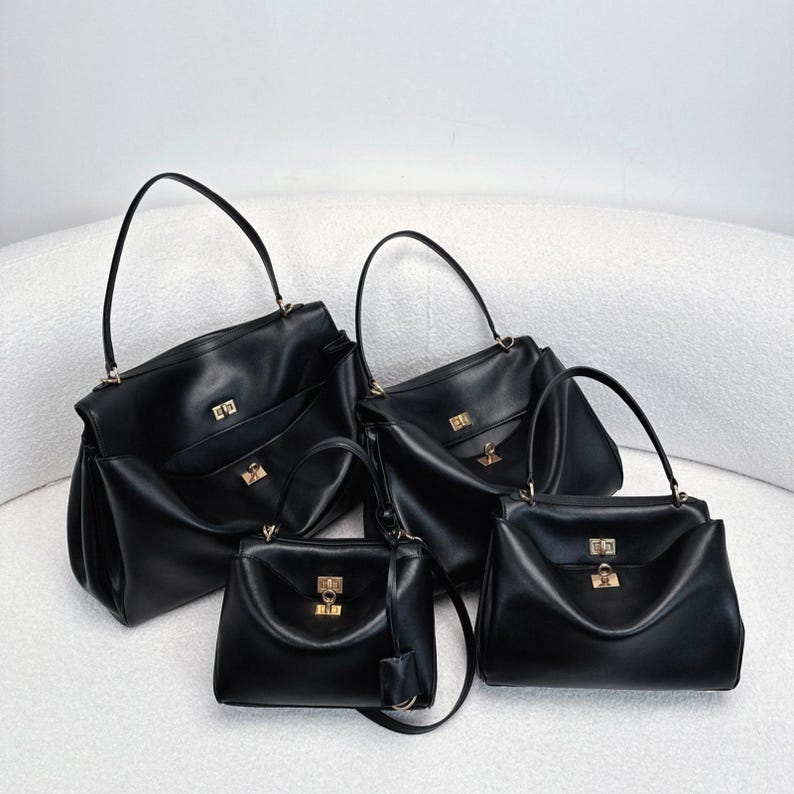 May include: Four black leather handbags of varying sizes are displayed. Each bag features a structured design with a top handle, a gold-colored clasp, and a front pocket. The bags are arranged on a white, curved surface.