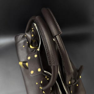 May include: A dark brown leather handbag with gold-colored studs and accents. The bag features two top handles and a zipper closure. The leather has a textured appearance, and the bag appears to be new.
