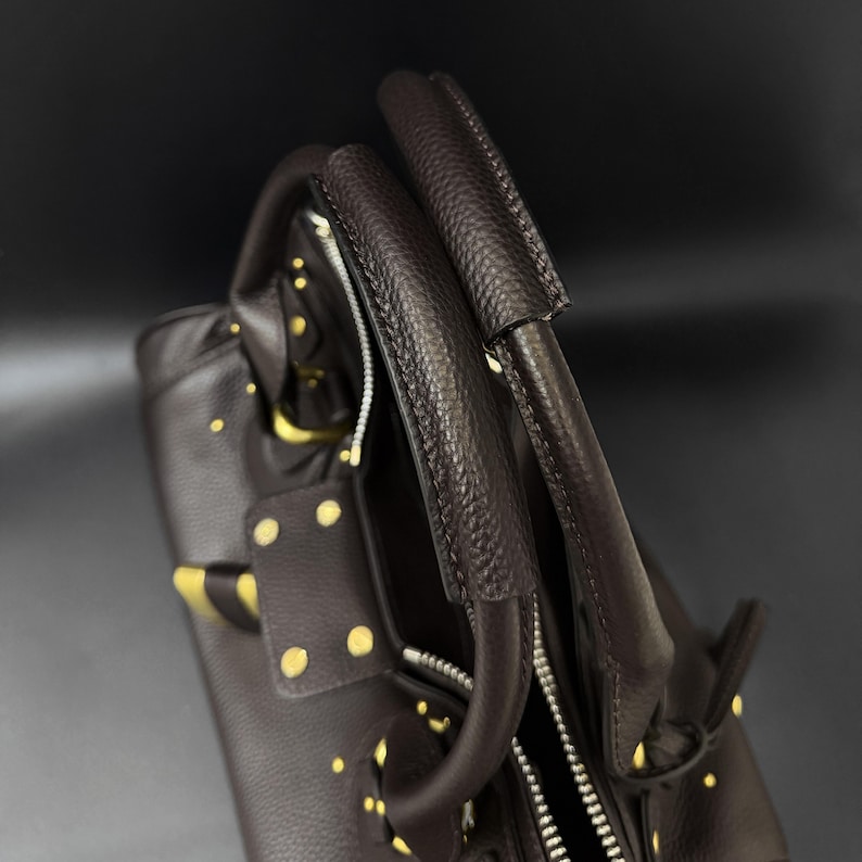 May include: A dark brown leather handbag with gold-colored studs and accents. The bag features two top handles and a zipper closure. The leather has a textured appearance, and the bag appears to be new.