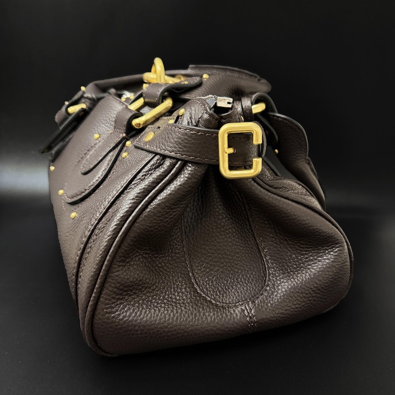 May include: A brown leather handbag with gold-colored hardware. The bag features a textured surface, a top handle, and a buckle closure. Small gold studs are visible on the exterior. The bag is set against a dark background.