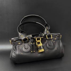 May include: Black leather handbag with gold-tone hardware, including a padlock closure and studded accents. Features dual top handles and a zippered opening. The bag has a structured shape and a pebbled texture.