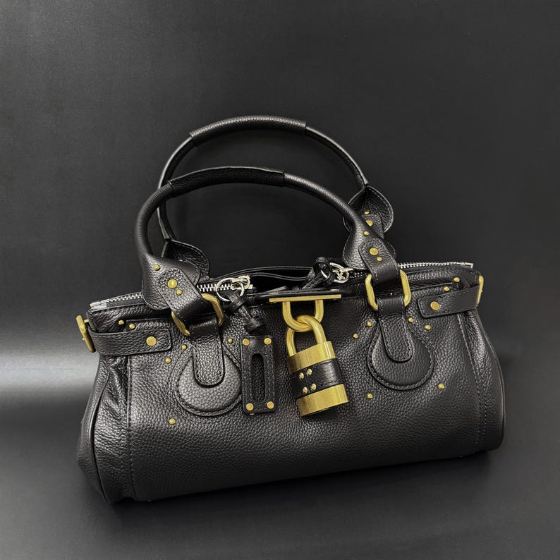 May include: Black leather handbag with gold-tone hardware, including a padlock closure and studded accents. Features dual top handles and a zippered opening. The bag has a structured shape and a pebbled texture.