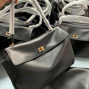 May include: Black leather handbags with top handles and flap closures. The bags have gold-colored hardware and are stacked together. The handles are wrapped in white paper. The bags are a classic style.