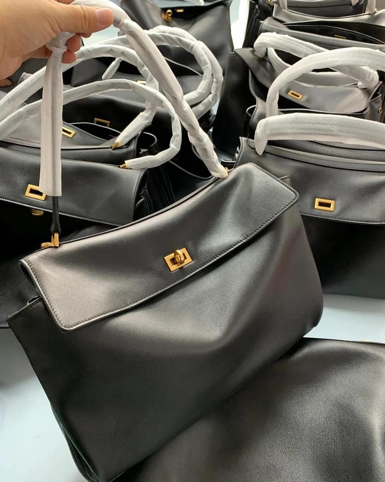 May include: Black leather handbags with top handles and flap closures. The bags have gold-colored hardware and are stacked together. The handles are wrapped in white paper. The bags are a classic style.