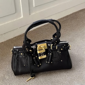 May include: Black leather handbag with gold-tone hardware, including a padlock closure and studded accents. Features a top handle and a key charm. The bag is sitting on a neutral-colored surface.