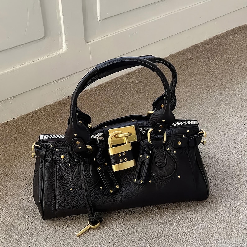 May include: Black leather handbag with gold-tone hardware, including a padlock closure and studded accents. Features a top handle and a key charm. The bag is sitting on a neutral-colored surface.