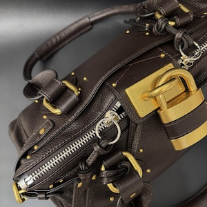 May include: A dark brown leather handbag with gold-toned hardware, including a padlock, zipper, and studs. The bag has a top handle and a textured surface, suggesting a high-end design. The zipper is silver-toned.