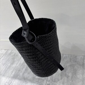 May include: A black woven leather bucket bag with a long strap. The bag has a braided design and a simple, minimalist style.