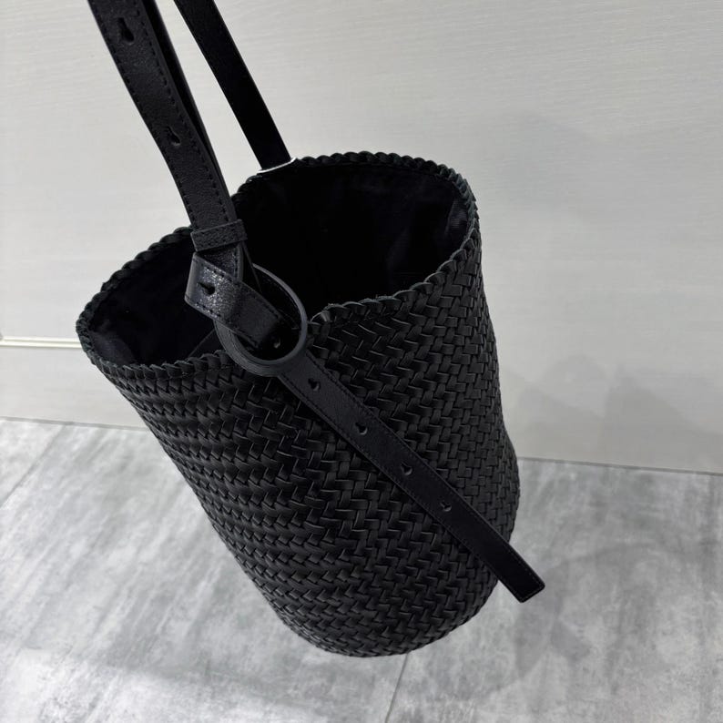 May include: A black woven leather bucket bag with a long strap. The bag has a braided design and a simple, minimalist style.