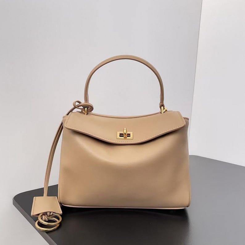 May include: A beige leather handbag with a top handle and a flap closure. The bag features gold-tone hardware and a detachable strap with a small tag and rings. The bag is sitting on a black surface.
