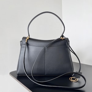 May include: Black leather handbag with a top handle and a detachable shoulder strap. The bag features gold-tone hardware and a structured design. The bag is sitting on a black surface.