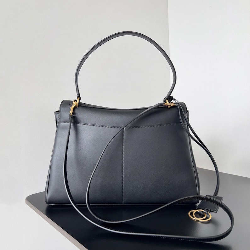 May include: Black leather handbag with a top handle and a detachable shoulder strap. The bag features gold-tone hardware and a structured design. The bag is sitting on a black surface.