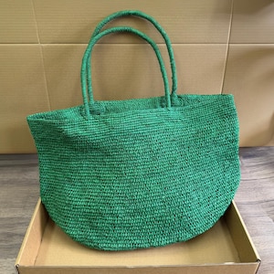 May include: A green woven tote bag with two matching handles. The bag has a rounded bottom and is made of a textured material. The bag is sitting in a cardboard box.
