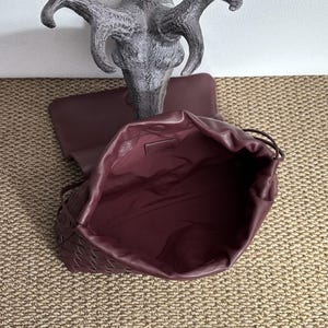 May include: A burgundy leather drawstring bag with a woven pattern on the front. The bag is open, revealing a spacious interior. The bag is set against a neutral background, with a decorative sculpture in the background.