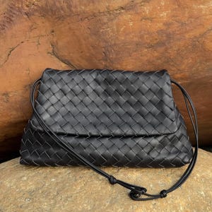 May include: A black woven leather clutch with a long, thin strap. The bag has a rectangular shape with a flap closure. The woven pattern is consistent across the entire surface of the bag.
