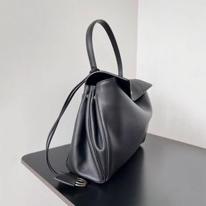 May include: Black leather handbag with a curved top handle and a flap closure. The bag has a structured design with a drawstring closure and a small, decorative tag with silver hardware. The bag is sitting on a black surface.