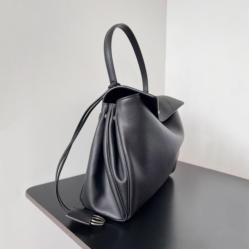 May include: Black leather handbag with a curved top handle and a flap closure. The bag has a structured design with a drawstring closure and a small, decorative tag with silver hardware. The bag is sitting on a black surface.
