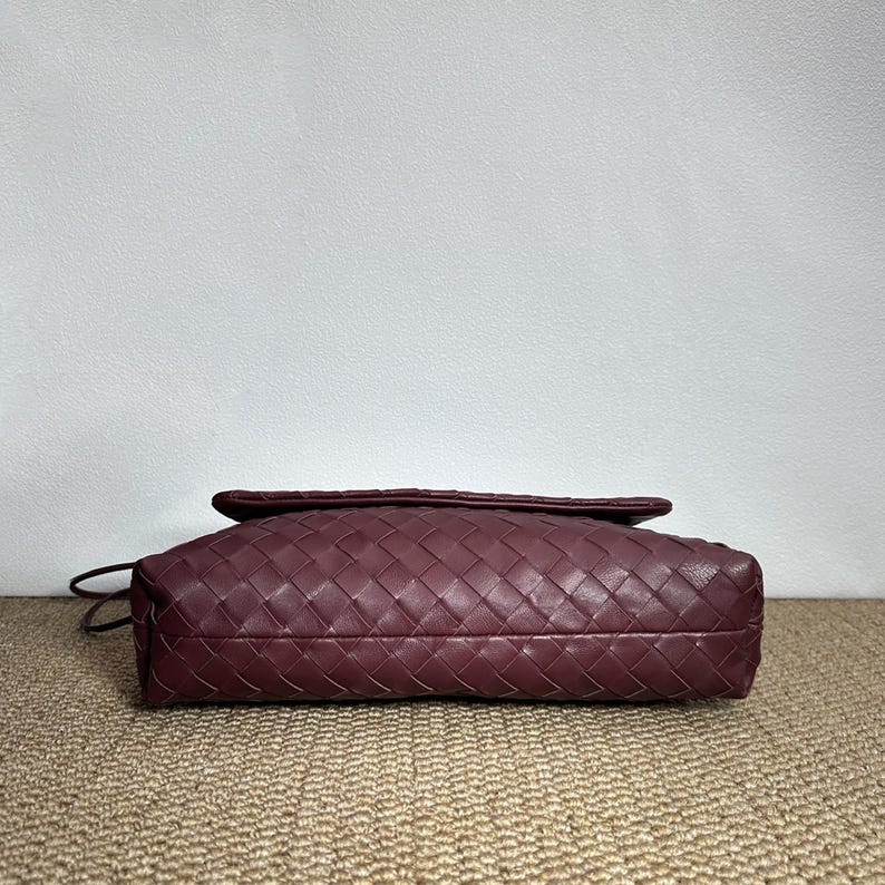 May include: A burgundy leather clutch with a woven design. The bag has a flap closure and a thin strap. The bag is rectangular and appears to be empty. The bag is sitting on a tan surface.