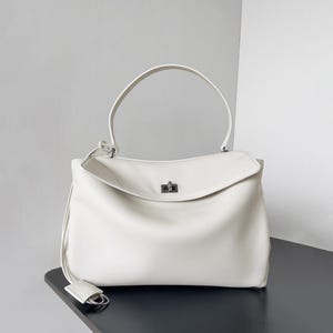May include: A cream-colored handbag with a top handle and a flap closure. The bag has a rectangular shape with a curved top and a small, silver-colored clasp. A long, thin strap with a small tag hangs from the side.