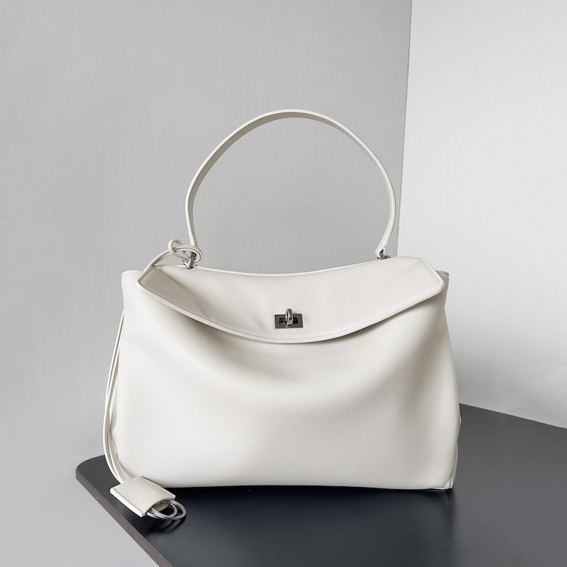 May include: A cream-colored handbag with a top handle and a flap closure. The bag has a rectangular shape with a curved top and a small, silver-colored clasp. A long, thin strap with a small tag hangs from the side.