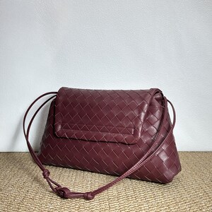 May include: A burgundy leather shoulder bag with a woven pattern. The bag has a flap closure and a long, thin strap. The bag is sitting on a tan surface.
