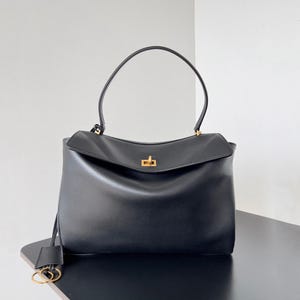 May include: Black leather handbag with a top handle and a flap closure. The bag features a gold-colored clasp and a small tag with gold-colored hardware. The bag is sitting on a black surface.