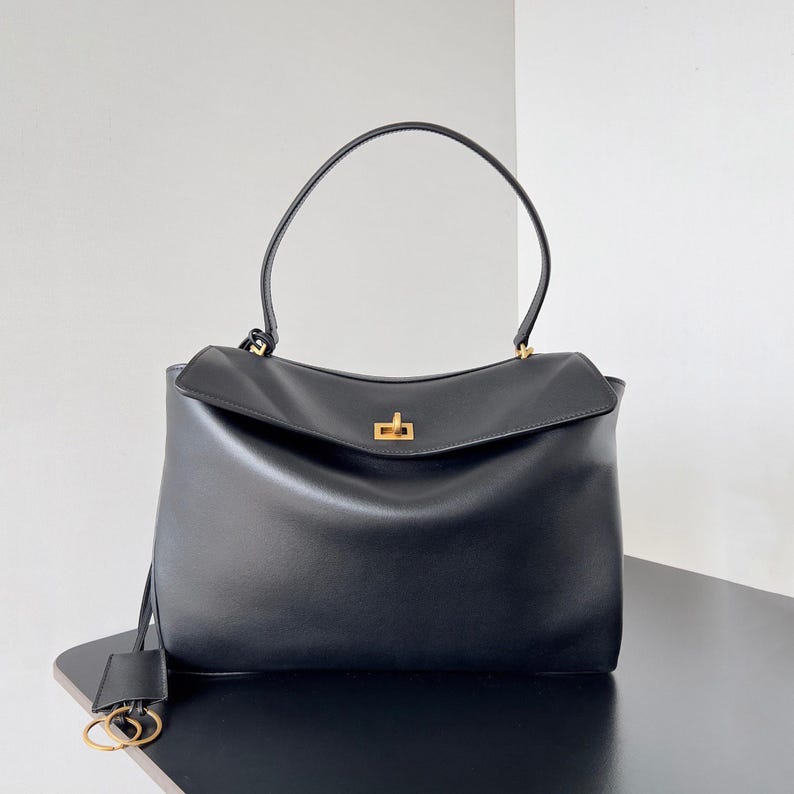 May include: Black leather handbag with a top handle and a flap closure. The bag features a gold-colored clasp and a small tag with gold-colored hardware. The bag is sitting on a black surface.