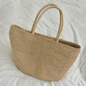 May include: A large, beige, woven straw tote bag with a wide, open-top design and two long, rounded handles. The bag features a consistent, textured, grid-like weave pattern throughout its entire structure, providing a sturdy and open appearance.