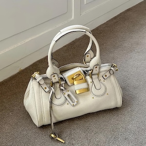 May include: Cream-colored handbag with a top handle and gold-tone hardware. The bag features a lock closure with a key and decorative studs. The bag is made of a textured material and has a structured shape.