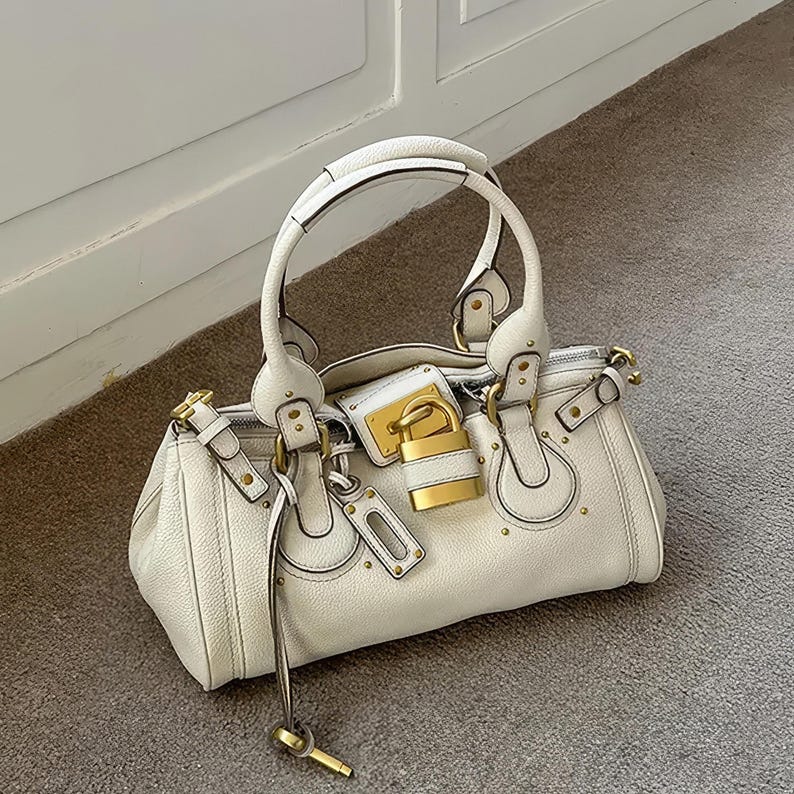 May include: Cream-colored handbag with a top handle and gold-tone hardware. The bag features a lock closure with a key and decorative studs. The bag is made of a textured material and has a structured shape.