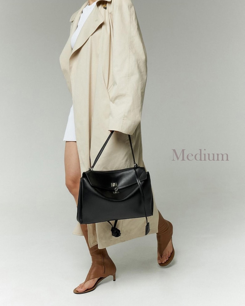 May include: A black leather handbag with a flap closure and a silver-tone clasp. The bag has a top handle and a long shoulder strap. The word "Medium" is visible in the image.