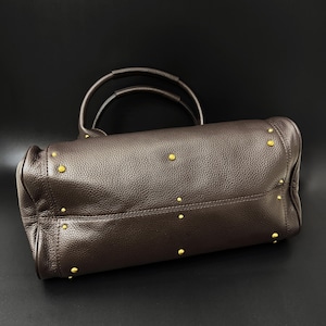 May include: A brown leather handbag with two handles and gold-colored studs. The bag has a textured surface and a structured shape, suitable for carrying essentials. The bag is set against a black background.