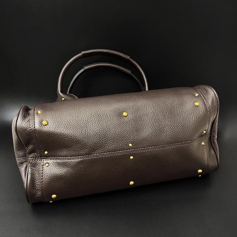 May include: A brown leather handbag with two handles and gold-colored studs. The bag has a textured surface and a structured shape, suitable for carrying essentials. The bag is set against a black background.