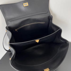 May include: Open black leather handbag with gold-toned hardware. The bag features a flap closure with a rectangular clasp, a top handle, and a zippered interior pocket. The interior is also black.