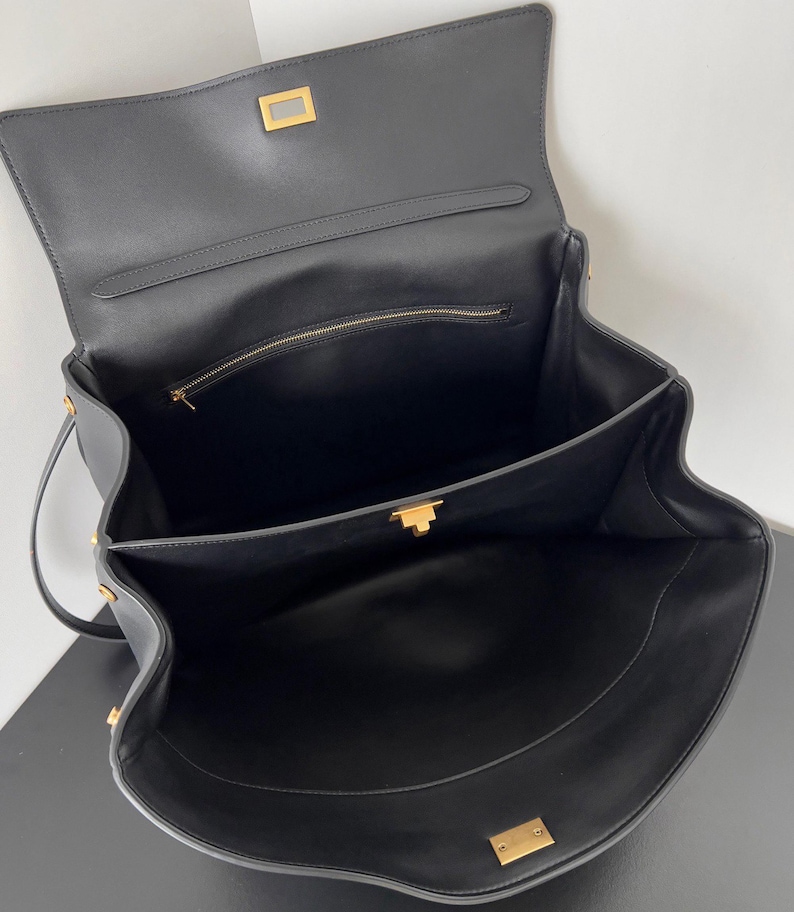May include: Open black leather handbag with gold-toned hardware. The bag features a flap closure with a rectangular clasp, a top handle, and a zippered interior pocket. The interior is also black.